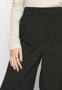 Black flowy trousers with an elastic waistband, paired with a light beige long-sleeve top. A gold ring is worn on the hand resting on the pant leg.