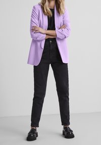 Kvinna står med armarna i kors iförd en lila kavaj, svart topp, svarta jeans och svarta platåskor framför en enkel ljus bakgrund.