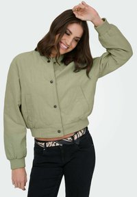 Veste bomber matelassée vert clair avec un col montant, des boutons-pression et des manches élastiques. Elle présente deux poches avant et une coupe courte.