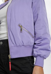 Lilāks bomber jakas ar rāvējslēdzēja krūšu kabatu, elastīgām piedurknēm un apakšmalu, ar sarkanu rāvējslēdzēja vilkšanas elementu un gludu, spīdīgu tekstūru.