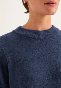 Navyblauer Pullover mit strukturiertem Strick, der über einen gerippten Rundhalsausschnitt und eine weiche, leicht fuzzy Oberfläche verfügt. Nahaufnahme einer Person, die ihn trägt.