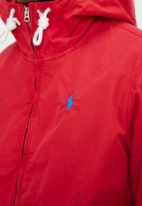 Czerwona kurtka z kapturem, wykonana z gładkiego materiału, z zapięciem na zamek z przodu i białymi sznurkami. Niebieskie logo na piersi.