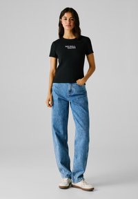Schwarzes, geripptes T-Shirt mit kurzen Ärmeln und "RED BULL RACING" in Weiß gedruckt. Kombiniert mit hellblauen, locker sitzenden Jeans und weißen Turnschuhen.