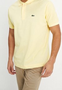 Žluté bavlněné polo tričko s klasickým límcem a zapínáním na tři knoflíky. Na levé hrudi se nachází zelené logo Lacoste.