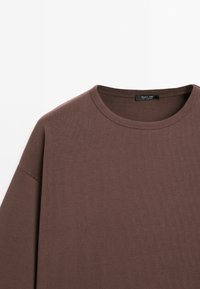 Sweat-shirt marron col rond avec une étiquette noire visible à l'intérieur du col, présenté sur fond blanc.