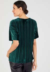 Top de terciopelo verde esmeralda con rayas verticales, mangas cortas y un escote redondo, con un corte holgado y una textura suave.