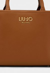 Hnedá kožená kabelka s textúrovaným povrchom a zlatým logom "LIU·JO MILANO" na prednej strane. Kompaktný dizajn s krátkymi uchami.