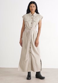 Μπεζ, χωρίς μανίκια μάξι φόρεμα με κουμπιά μπροστά, δύο τσέπες στο στήθος και δεμένο στη μέση. Μαλακό ύφασμα με ελαφριά γυαλάδα, φορεμένο με μαύρες μπότες.