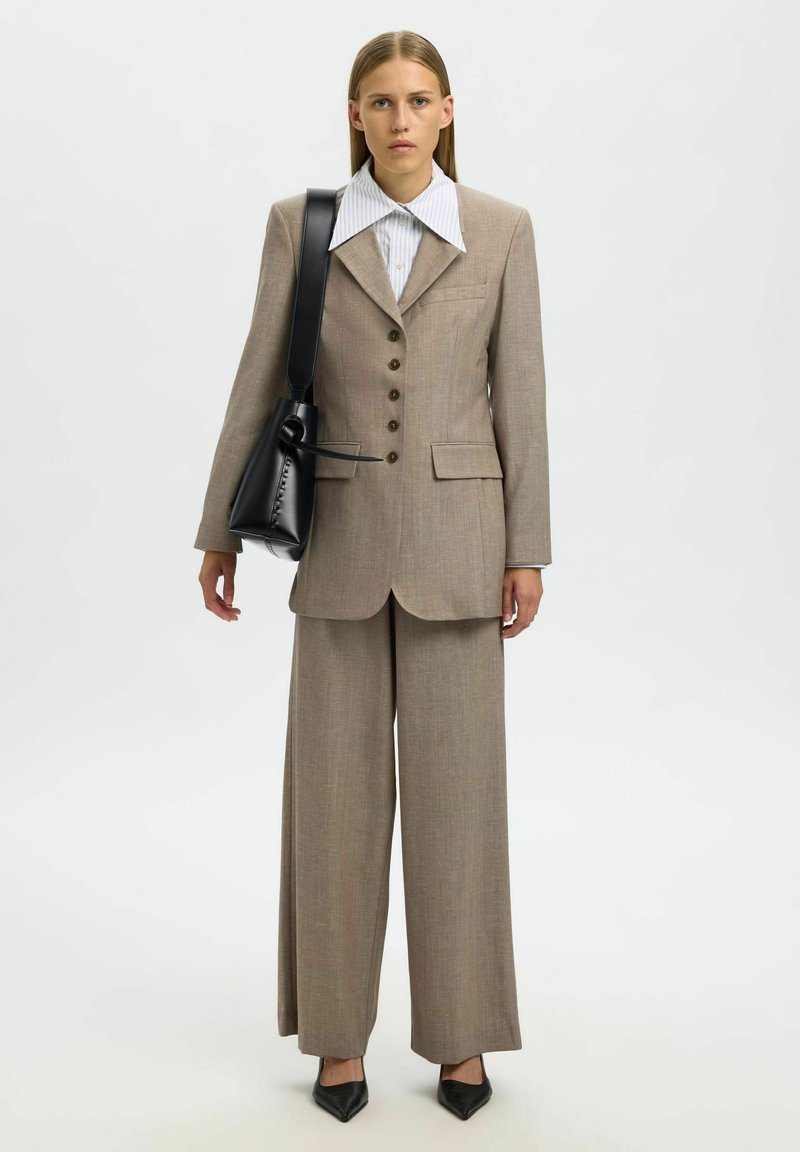 Femme portant un costume beige avec un pantalon à jambes larges, une chemise blanche à col, des chaussures noires pointues et un sac à épaule noir.