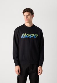 Young man wearing black sweatshirt with blue and green flame-style "HUGO" text, standing against a plain light gray background.