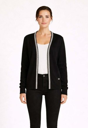 Donna che indossa un cardigan nero con rifiniture bianche sopra una maglietta bianca e pantaloni neri, in piedi davanti a uno sfondo chiaro e semplice.