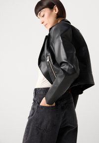 Zwart leren cropped jack met een ritszakdetail, gedragen over een lichtgekleurde top, gecombineerd met hoge zwarte denim jeans.