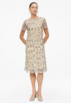 Frau trägt ein knieumspieltes beiges und braunes Kleid mit Zickzackmuster und beige Absatzsandalen, steht mit den Händen hinter dem Rücken.