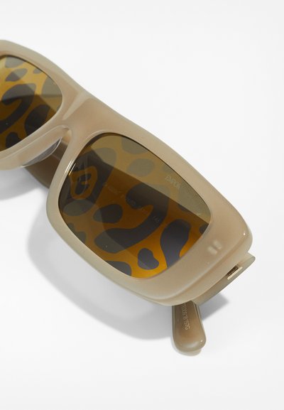 Lunettes de soleil oversize beiges avec une finition brillante, dotées de verres teintés marron avec un motif léopard et de charnières latérales pour un maintien sécurisé.