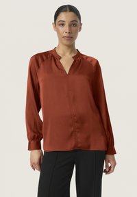Blusa color ruggine realizzata in tessuto morbido, con scollatura a V, maniche lunghe e sottili pieghe verticali lungo la parte anteriore.