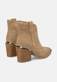 Botas de tobillo de ante en color beige con puntera redondeada, costuras decorativas y un tacón de madera con un acento plateado. Cierre con cremallera en la parte posterior.