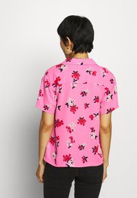 Kortärmad skjorta i rosa tyg med blommönster, med röda och vita blommor samt gröna blad, krage och detalj på ryggen.