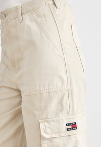 Beige cargo-housut, joissa on useita taskuja, näkyvät ompeleet, vetoketjuhihna ja sivutaskussa logo-patch, jossa lukee "TOMMY JEANS".