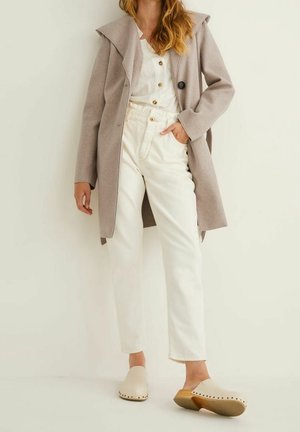 Woman wearing beige coat, white buttoned blouse, high-waisted white pants, and light-colored slip-on clogs, standing against a plain background.