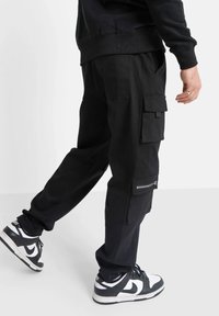 Pantaloni cargo neri con molte tasche, dettaglio con zip e vestibilità rilassata, abbinati a sneakers nere e bianche.