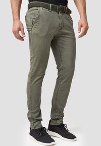 Pantalones khaki de corte slim con cinco bolsillos, un cinturón trenzado verde y una camiseta oscura. Combinados con zapatillas negras con suelas blancas.