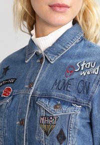 Blonďatá žena v modré džínové bundě s nášivkami s nápisy "Zůstaňte zdraví", "POJĎTE DAL" a "CO?" a s tváří mimozemšťana, přes bílý rolák.