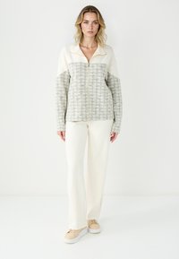 Veste zippée avec un corps beige clair texturé et des manches crème, associée à un pantalon crème et des baskets beiges, le tout sur fond blanc.