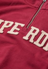 Bordeaux ritsweater met grote, beige geborduurde letters. Gladde, katoenmixstof met een halflange ritskraag.