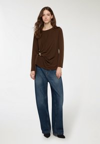 Maglia marrone a maniche lunghe con dettaglio annodato e hardware dorato all'orlo, abbinata a jeans blue a gamba larga, indossata con scarpe nere.