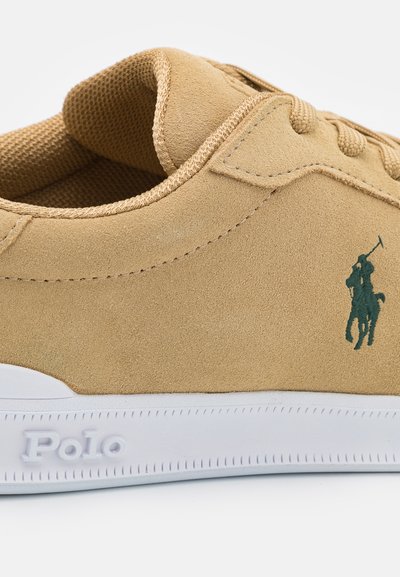 Sapatilhas de camurça em tom bege com um acabamento texturizado, apresentando bordado do logótipo em verde e sola de borracha branca. Ilhós estruturados para os atacadores.