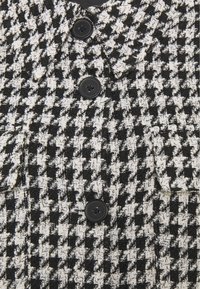 Kangas, jossa on musta-valkoinen hounds-tooth-kuvio, jolla on teksturoitu pinta. Sisältää kaksi mustaa nappia ja viilto-kauluksen. Minimalistinen taskuyksityiskohta.