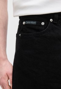 Κοντινή λήψη τσέπης μαύρου τζιν με ασημί μεταλλικά στερεώματα και ετικέτα Calvin Klein, φορεμένη με λευκό τοπ.