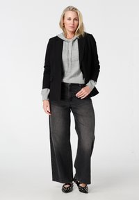 Zwarte blazer over een lichtgrijze hoodie, gecombineerd met high-waisted, wijde zwarte jeans. Gestructureerde zwarte schoenen met gouden accenten. Eenvoudige sieraden.