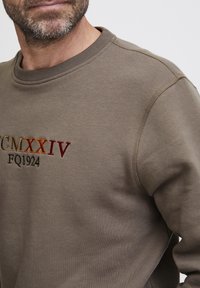 Sudadera marrón con cuello de canalé. Presenta un texto bordado multicolor: "CMXXIV" en tonos de naranja y rojo, y "FQ1924" en negro.