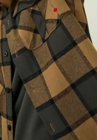 Manica di camicia a quadri marroni e neri con bottoni e asole nere, ripiegata su un indumento verde scuro sottostante.