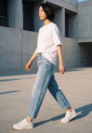 Jonge vrouw met kort zwart haar die buiten loopt, draagt een wit T-shirt, lichtblauwe gescheurde spijkerbroek en witte sneakers.