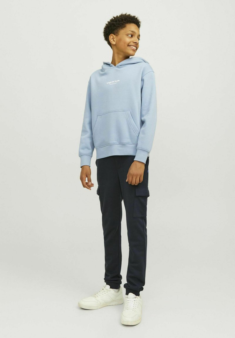 Felpa azzurra chiara con tasca a marsupio e logo bianco, abbinata a pantaloni cargo scuri con tasche laterali e sneakers bianche.