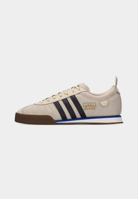 Beige Adidas Samba -tossu mustilla raidoilla, sinivalkoisella välipohjalla, ruskealla ulkopohjalla ja kultaisella logolla sivussa.