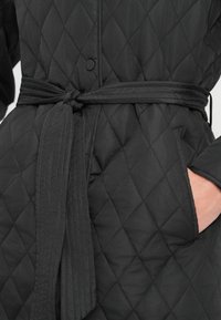 Μαύρο παλτό με καπιτονέ ραφές που διαθέτει ζώνη στη μέση, διαμάντι σχέδιο, τσέπες με καπάκι και κούμπωμα με κουμπιά. Η υφή του υφάσματος προσθέτει βάθος στο σχέδιο.