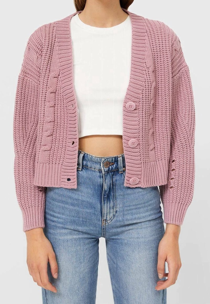 Femme portant un cardigan rose tricoté court à boutons sur un top blanc court et un jean bleu taille haute.