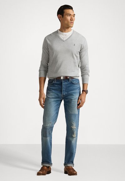 Suéter cinza com decote em V, t-shirt branca clara por baixo, jeans azuis desfiados com remendos, sapatos de couro castanho, relógio de metal, pose em pé.