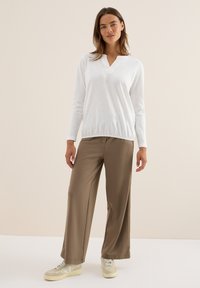 Femme debout portant un haut blanc à manches longues en V, un pantalon large marron et des baskets beiges, sur un fond beige uni.