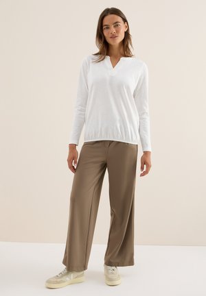 Kvinde står iført en hvid langærmet V-hals top, brede brune bukser og beige sneakers foran en ensfarvet beige baggrund.