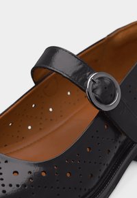 Chaussure Mary Jane en cuir verni noir avec un design perforé et une bride à boucle ronde argentée, doublure intérieure beige visible.