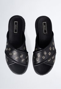 Sandalias negras sin hebilla con correas entrelazadas adornadas con pequeños logotipos metálicos y suelas acolchadas.