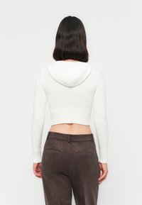 Person mit schulterlangen, dunklen Haaren, die einen weißen, kurz geschnittenen Hoodie und braune Cordhosen trägt, mit dem Rücken zu einem schlichten weißen Hintergrund.