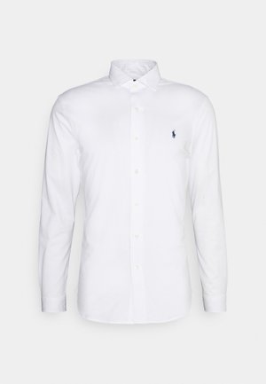 Vit långärmad skjorta med knappar i bomull. Har en spridningskrage, framknäppning och en liten marinblå logotyp på vänster bröst.