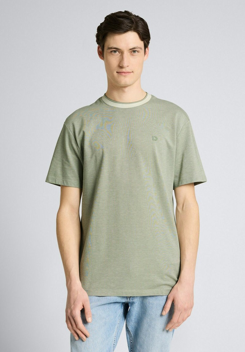 Junger Mann, der nach vorne blickt, trägt ein hellgrünes kurzärmliges T-Shirt und hellblaue Jeans vor einem einfarbigen Hintergrund.