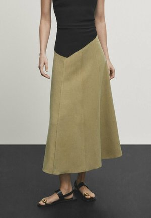 Olijfgroene A-lijn rok met een zwarte strak zittende top met een asymmetrische halslijn. Platte sandalen met een cirkelvormige metalen accent.