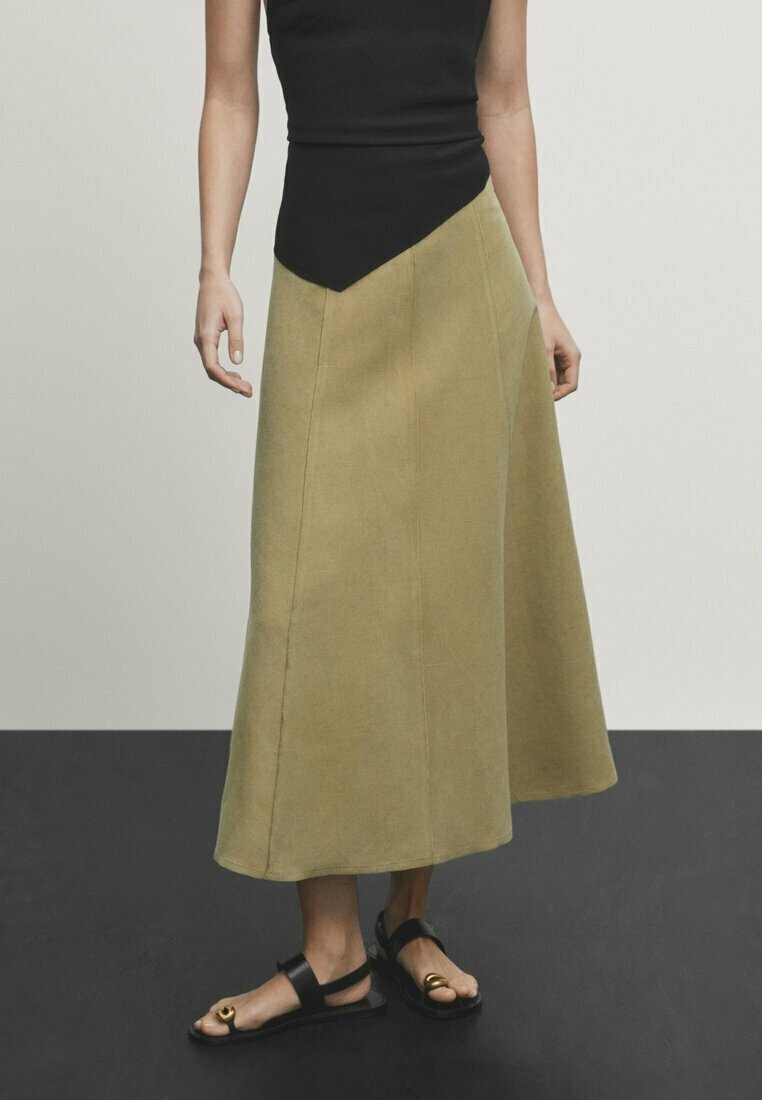 Jupe trapèze vert olive avec un haut ajusté noir, présentant un décolleté asymétrique. Sandales plates avec un accent métallique circulaire.